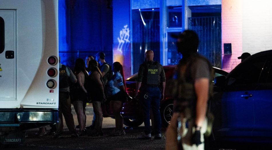Un agente de HSI custodia a un grupo de personas en fila junto a un autobús y vehículos durante el operativo de ICE en El Corralón, con un agente adicional con el rostro cubierto en primer plano.